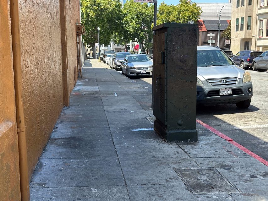 A sidewalk with a large utility box obstructing the path, alongside parked cars and buildings in an urban area on a sunny day.