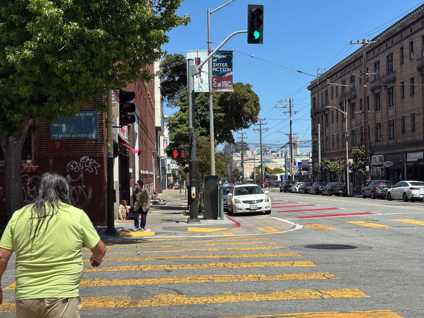 A person with long gray hair crosses a city street at a crosswalk; the traffic light is green and a white car waits at the intersection.