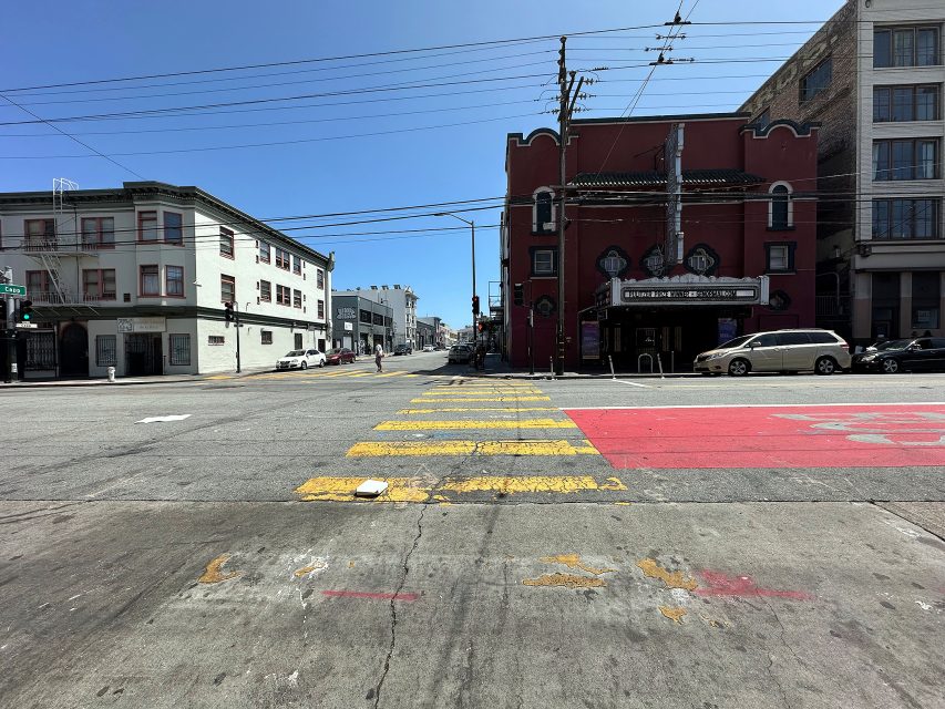 A city intersection with yellow crosswalk lines, a red building on the right, white buildings on the left, and clear blue sky overhead.