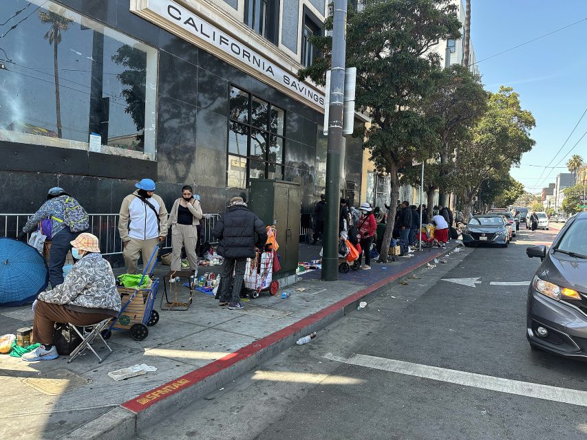People stand and sit with belongings along a city sidewalk in front of a building labeled "California Savings." Some items and carts are scattered along the curb.