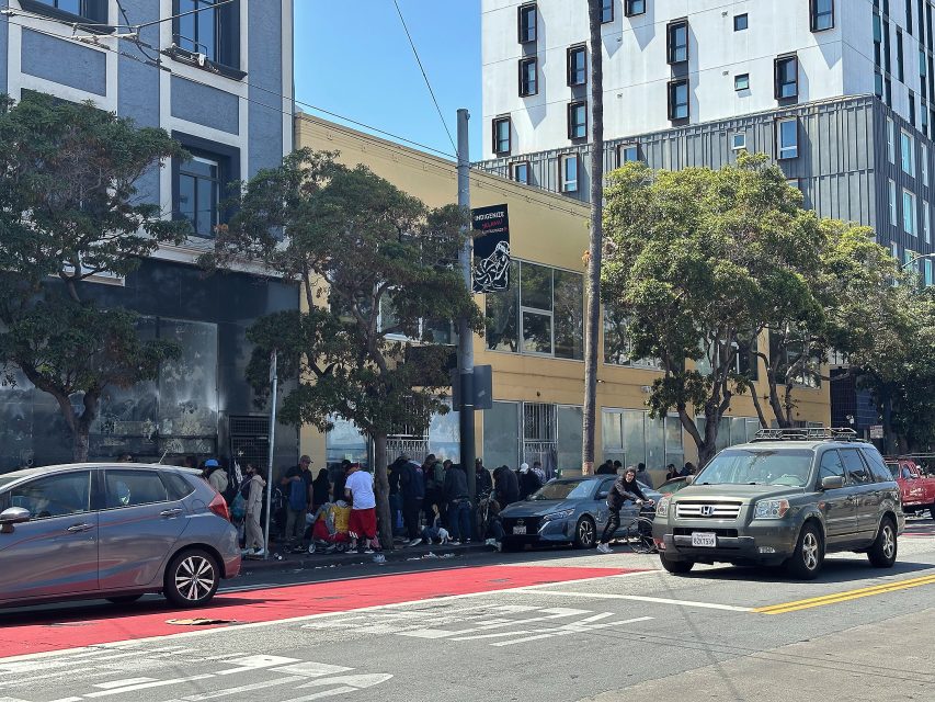 A crowd of people stands in line on a city sidewalk beside parked cars and buildings on a sunny day.