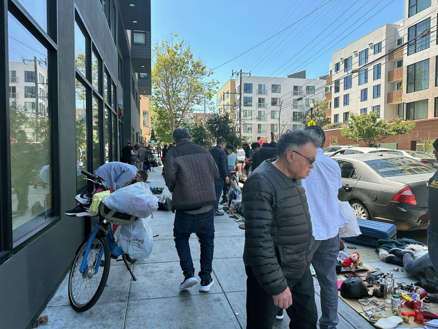 People browse items laid out for sale on a city sidewalk; some belongings are attached to a bicycle, and buildings line the background.