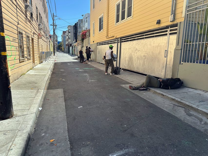 A narrow urban alley with several people; one person lies on the ground while others stand or walk along the sidewalk and street.