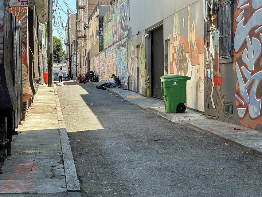 A city alleyway with graffiti-covered walls, a green trash bin, and a few people sitting or lying on the ground in the distance.