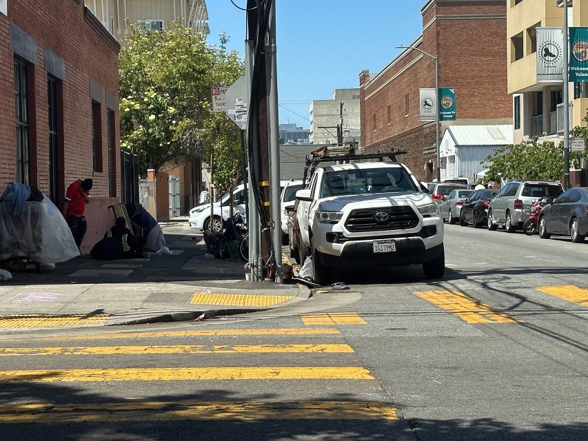 A white pickup truck is parked beside a yellow crosswalk on a city street. Several tents and people are visible on the sidewalk to the left.