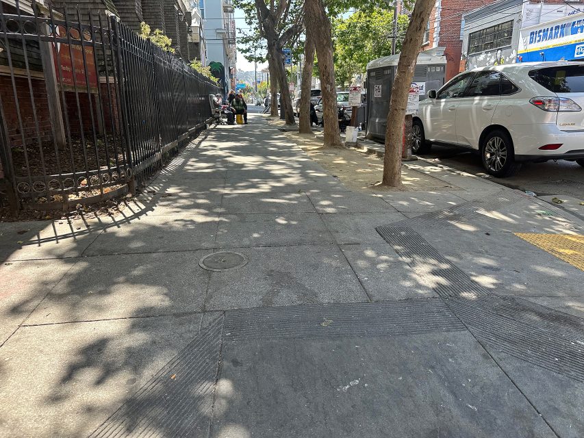A city sidewalk lined with trees and parked cars, featuring a metal fence on the left and a utility box on the right; sunlight creates dappled shadows on the pavement.