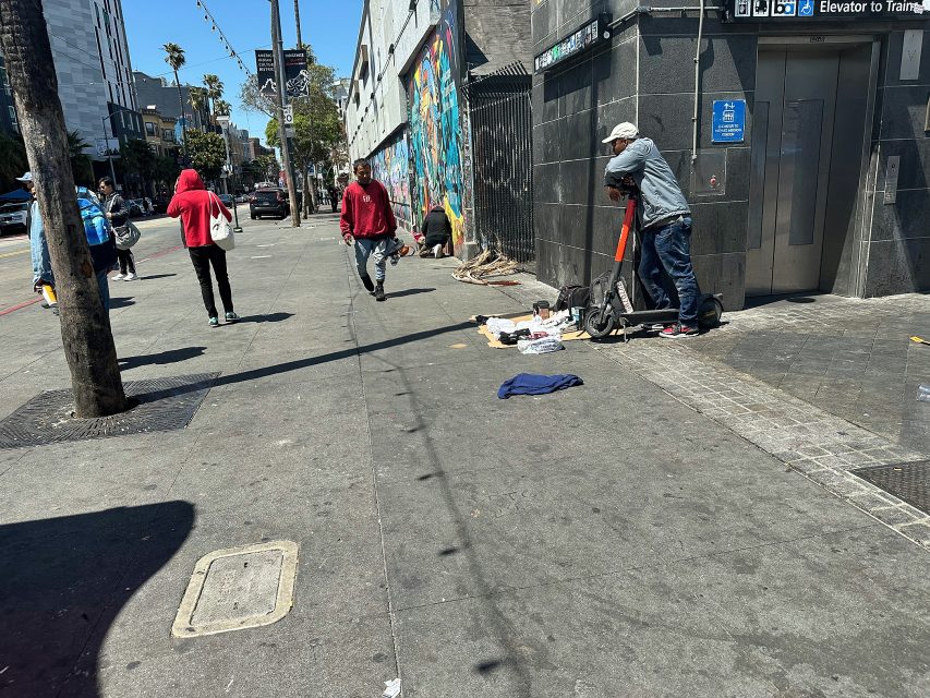 A man leans on a scooter near an elevator entrance as others walk by on a city sidewalk scattered with belongings and clothing during the day.