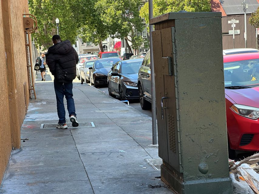 A person in a black jacket walks down a city sidewalk lined with parked cars and trees, with a utility box in the foreground.