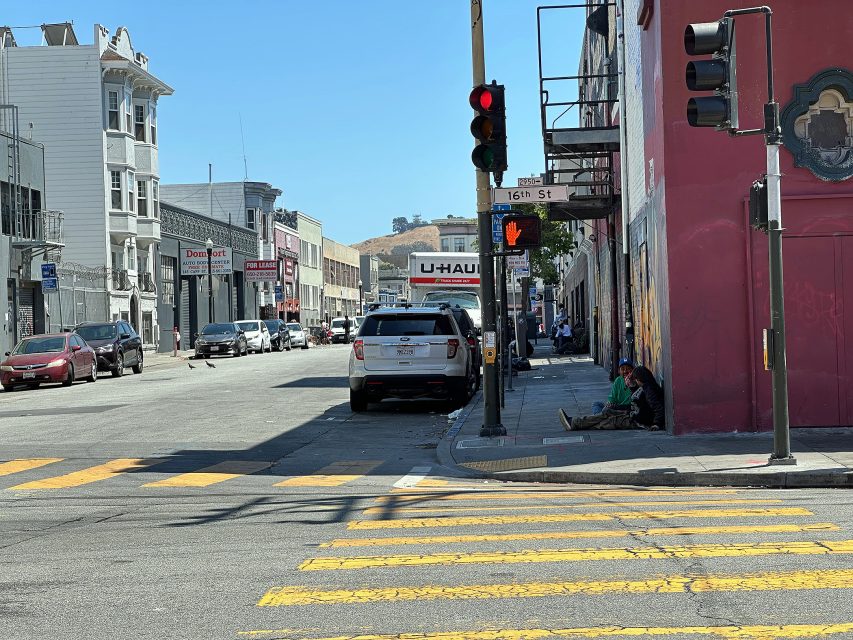 Street scene at a red light on 16th St; parked cars, a U-Haul sign, pedestrians on the sidewalk, and a few people sitting near a building with a red wall.