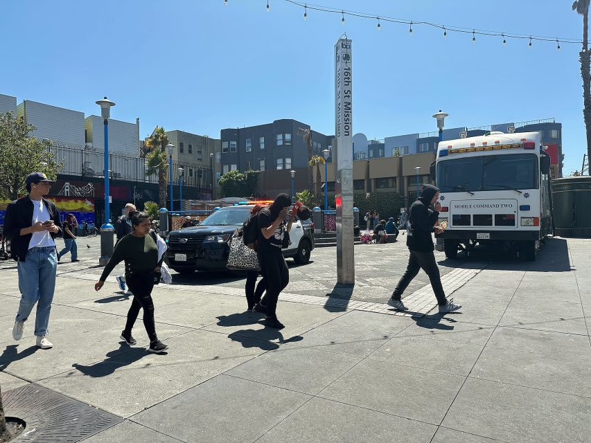 People walk past a parked police car and a mobile command van in a sunny urban plaza, with buildings and string lights in the background.