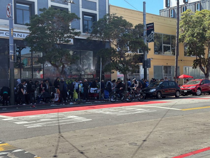 A long line of people stands on a city sidewalk near a building and trees, with several parked cars and bicycles along the street.