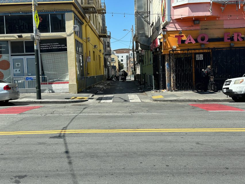A narrow alleyway between two buildings, with a person standing near a taqueria on the right and parked cars on both sides of the street in daylight.