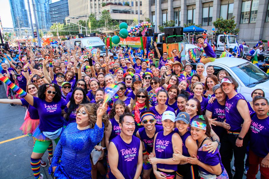 A large, diverse group of people in matching purple shirts pose and smile together at an outdoor Pride parade with colorful rainbow decorations.