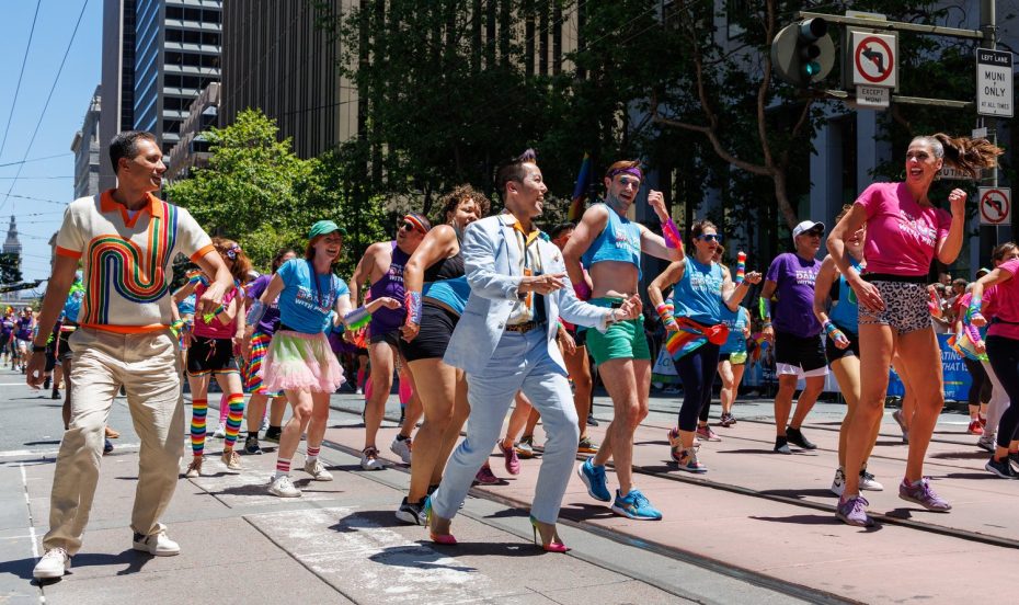 A group of people in colorful outfits dance and march down a city street during a parade on a sunny day.