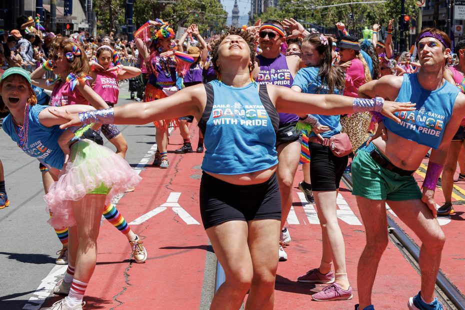 A group of people in colorful outfits dance on a city street during a pride parade, wearing blue shirts that say "Dance with Pride.