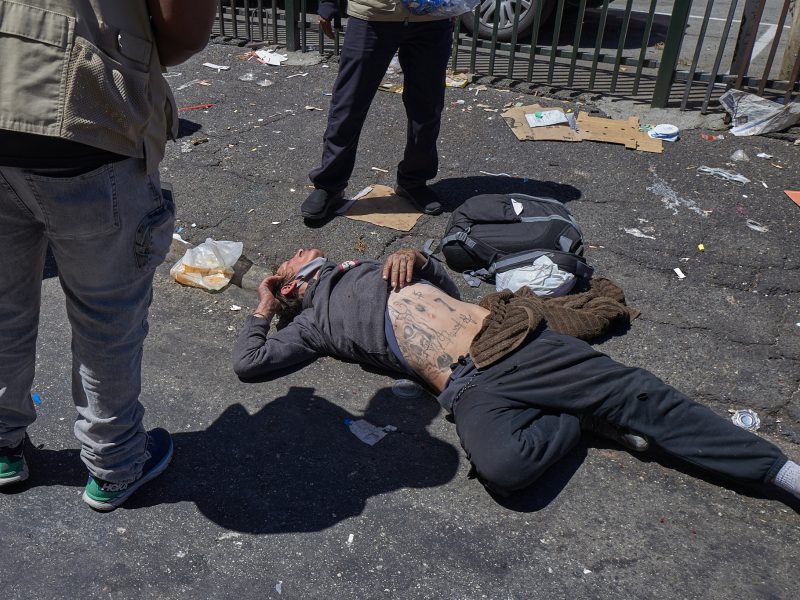 A person lies on a littered street with two people standing nearby. The person on the ground has tattoos on their torso and is surrounded by scattered belongings and debris.