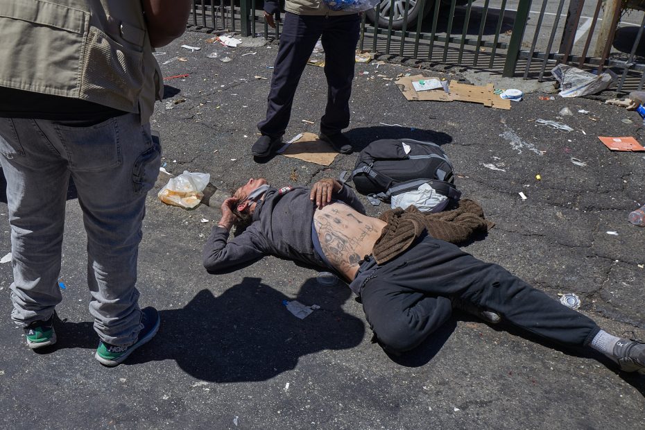 A person lies on a littered street with two people standing nearby. The person on the ground has tattoos on their torso and is surrounded by scattered belongings and debris.