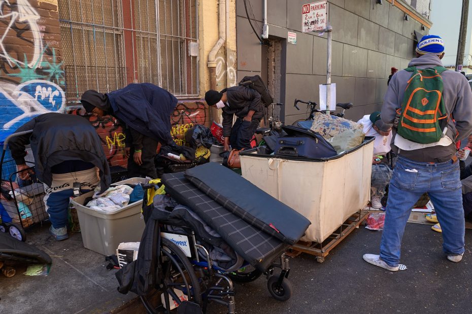 Several people sort through belongings and carts filled with items on a city sidewalk next to a graffiti-covered wall and building.