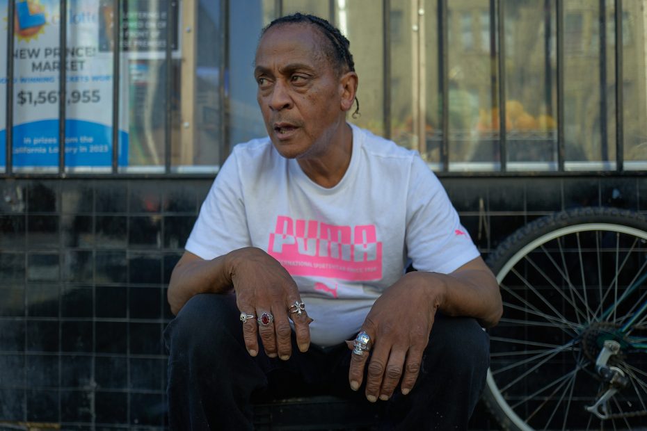 A person with braided hair wearing a white Puma shirt and multiple rings sits outside near a shop window and a bicycle wheel.