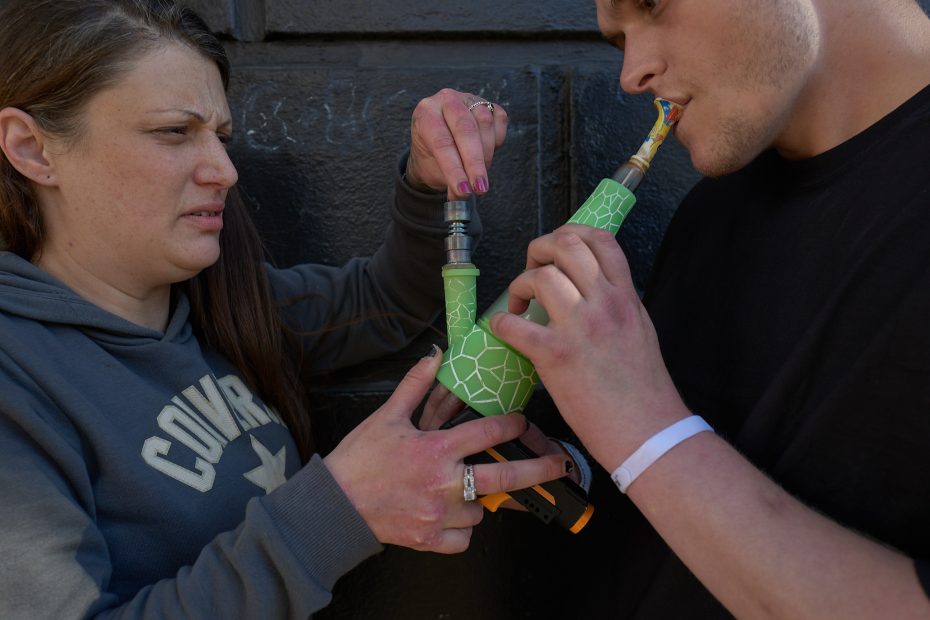 A woman hands a green, giraffe-patterned bong to a man as he lights it, while she looks on with a displeased expression.