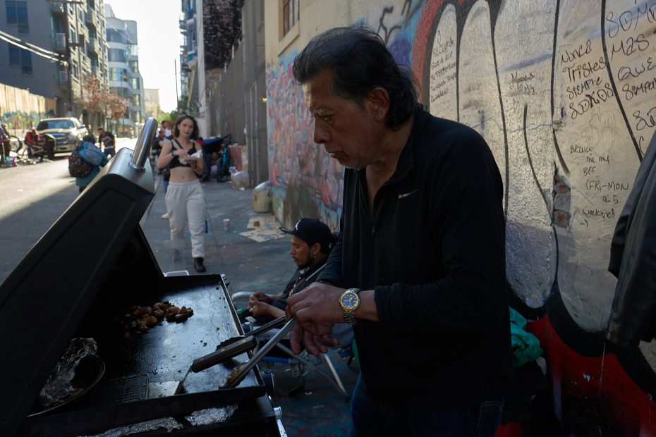 A man grills food on a barbecue in an urban alleyway with graffiti-covered walls; people stand and sit nearby.