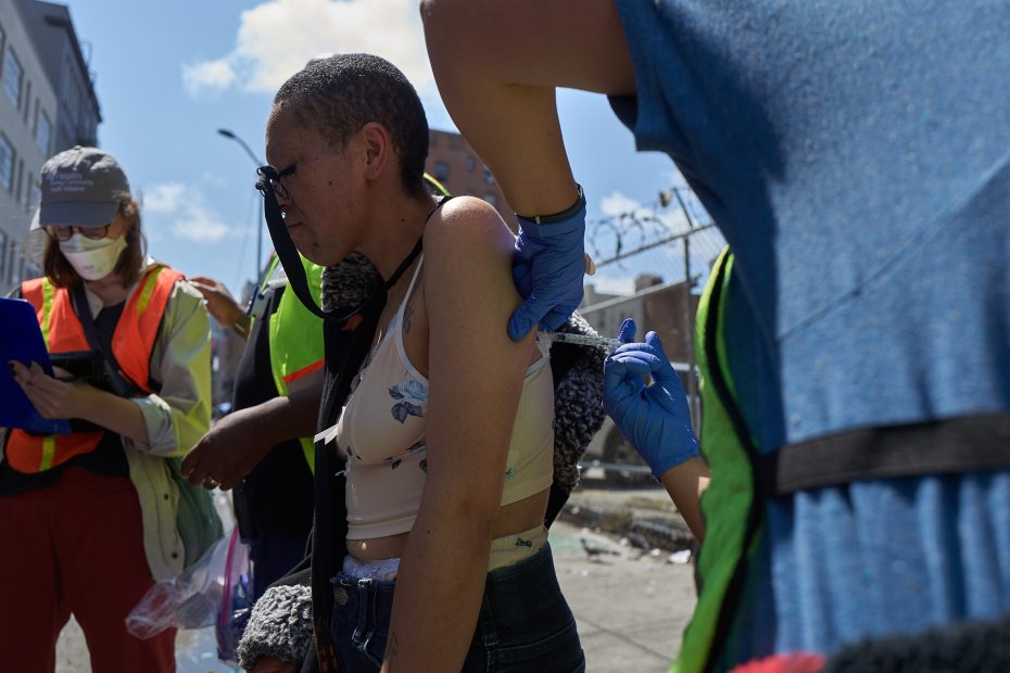 A person receives an injection in their upper arm outdoors, with others nearby wearing safety vests and masks under a partly cloudy sky.