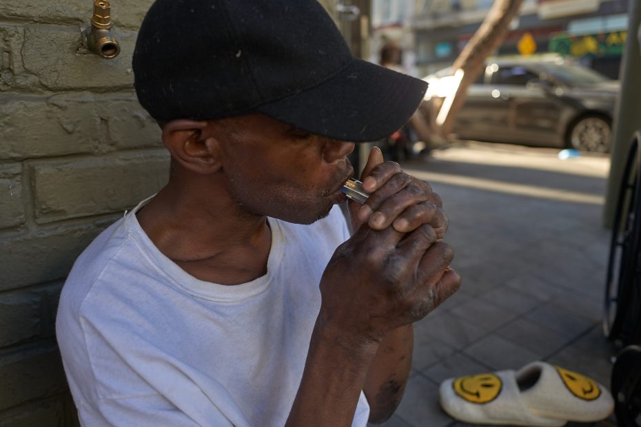 A man in a white t-shirt and black cap sits on a sidewalk, lighting a pipe. A pair of slippers with smiley faces are on the ground nearby.