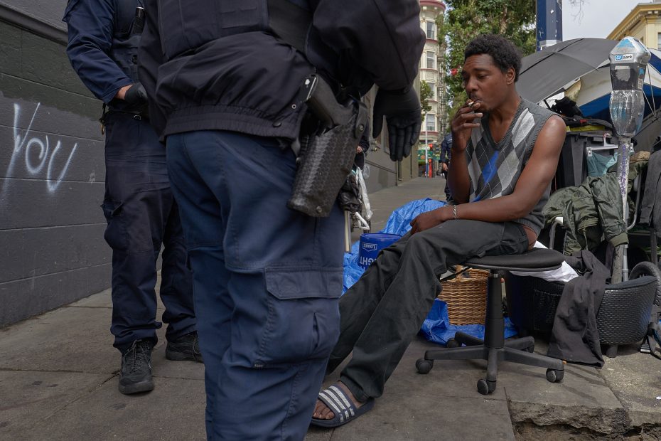 A man sits on a rolling chair on the sidewalk, smoking, while two uniformed police officers stand nearby; personal belongings are visible behind him.