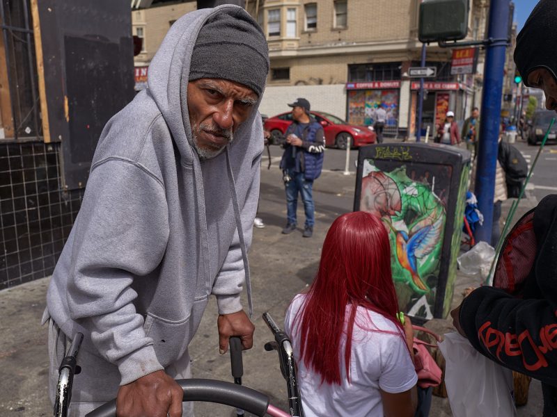 An older man in a gray hoodie leans on a walker on a city street, while a woman with red hair sits nearby and others stand around in the background.