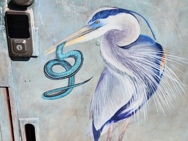 A painted image of a heron holding a blue snake in its beak is depicted on a door with a lock and handle visible.