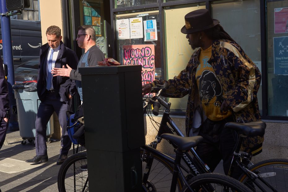 Three men stand on a city sidewalk near a window; one pushes a bicycle, while the others converse beside a utility box. Sunlight casts strong shadows.