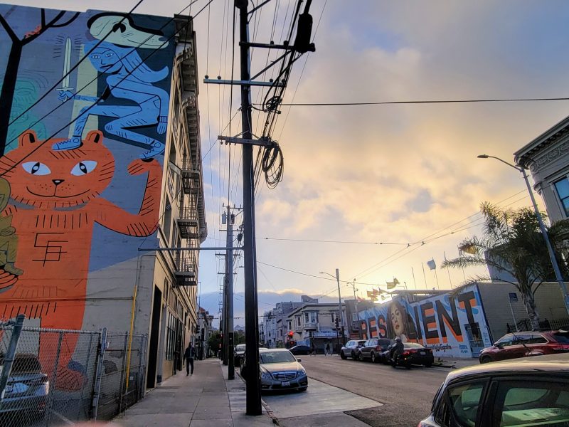 Street view with large colorful murals on building walls, parked cars along the sidewalk, power lines overhead, and a partly cloudy sky at sunset.