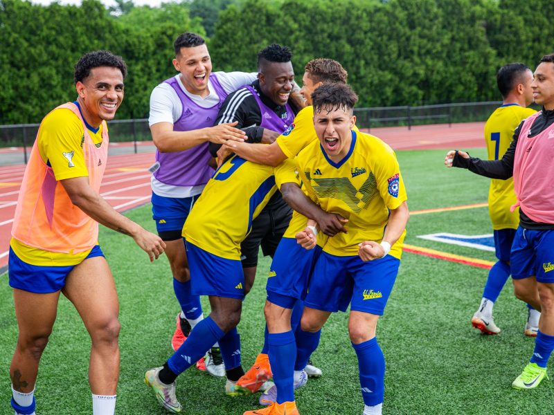 A group of soccer players in yellow and blue uniforms celebrate together on a field, displaying excitement and smiling.