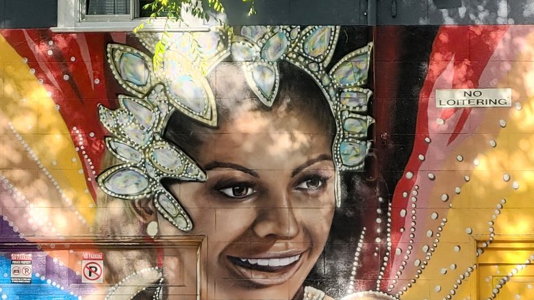A mural of a smiling woman wearing an elaborate jeweled headdress is painted on a wall with a "No Loitering" sign above her. Sunlight and leaves cast shadows on the artwork.