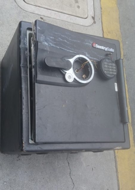 A damaged SentrySafe safe with a broken handle and exposed wires sits on a sidewalk near a yellow curb.