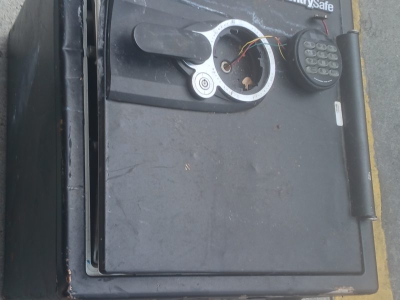 A damaged black SentrySafe safe lies on its side on a sidewalk near a yellow curb and a utility cover.