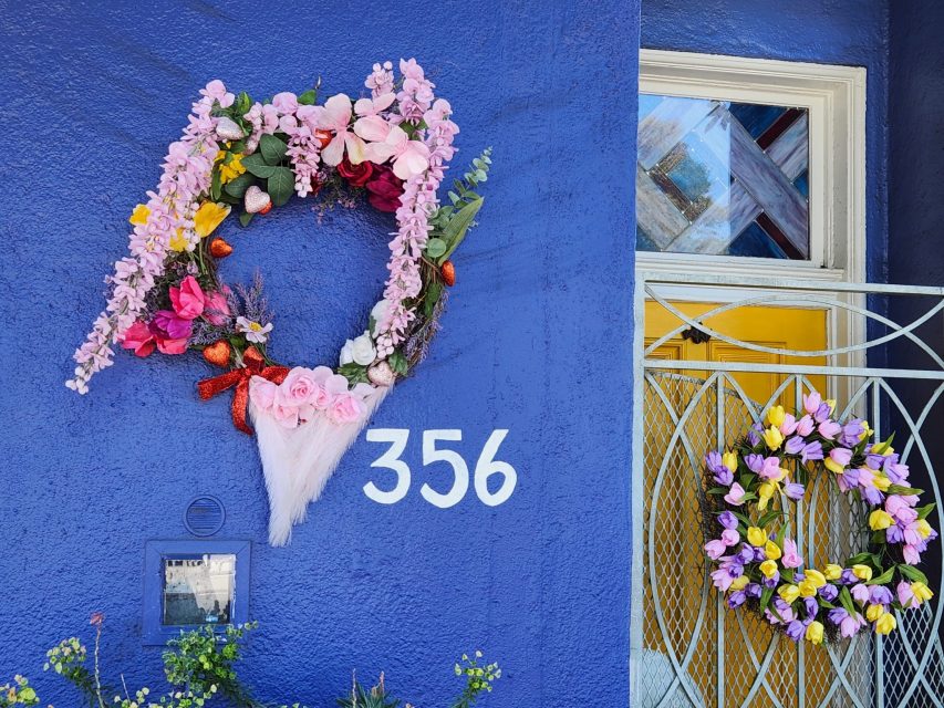 Two floral wreaths on a blue wall; one wreath in a heart shape above the number 356, and another circular wreath on a yellow door with a metal grill.