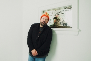 A man in a black sweater and orange beanie leans against a white wall beneath a window with a blurry reflection of himself.
