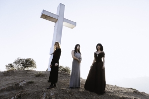 Three women in long dresses stand on a hillside near a large white cross, with sparse vegetation and a bright sky in the background.