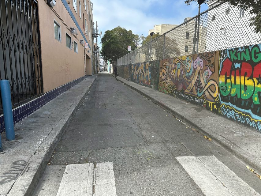 A narrow urban alleyway with graffiti-covered walls, a crosswalk in the foreground, and apartment buildings lining one side under a partly cloudy sky.