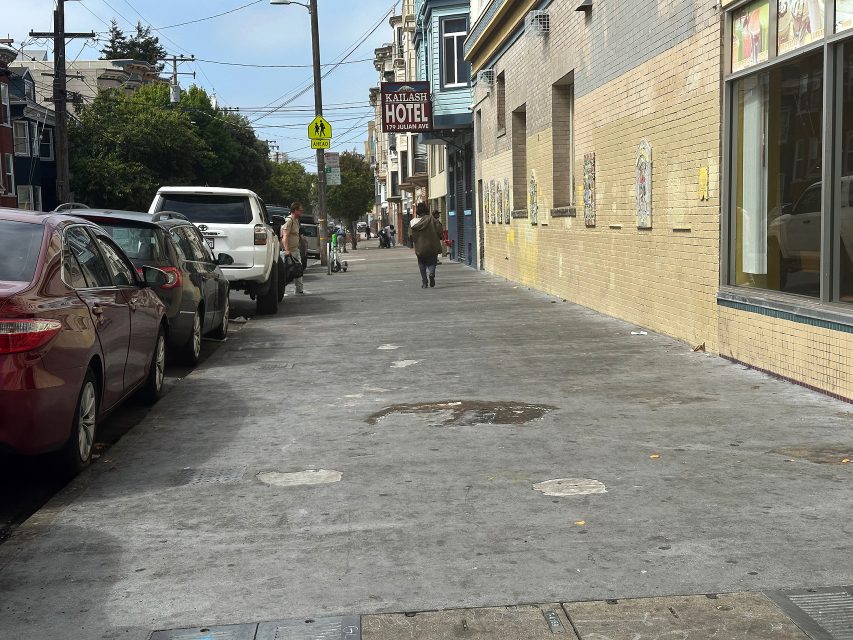 A city sidewalk with parked cars along the street, a person walking, and a sign for Kailash Hotel on a beige brick building.