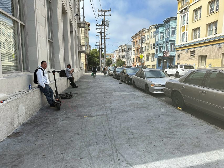 Two men stand on a wide city sidewalk next to a row of parked cars, with colorful buildings lining the street under a partly cloudy sky.