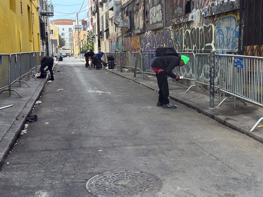 Several people clean a graffiti-covered alleyway behind metal barricades on an overcast day.