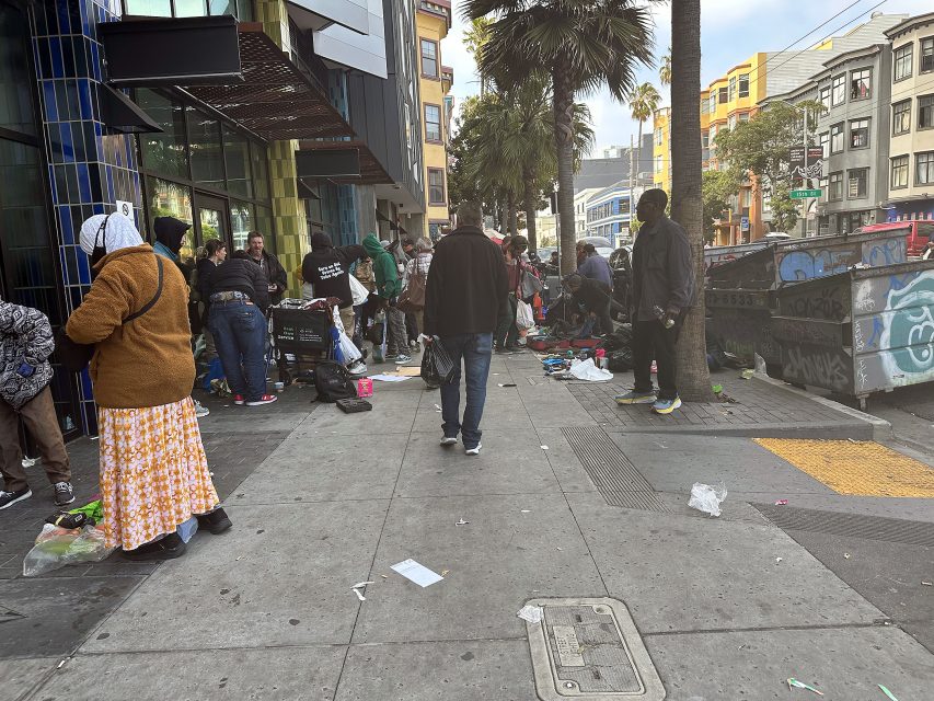 People gather on a city sidewalk with scattered belongings, near a building and palm tree, while others walk by; urban street scene with graffiti and litter.