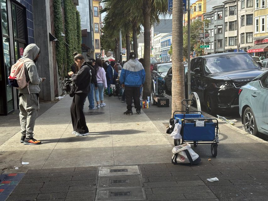 People stand and walk on a city sidewalk lined with parked cars and palm trees; a cart with bags is on the right side of the image.