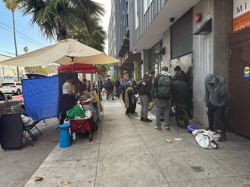 People stand and sit along a city sidewalk beside a building; a street vendor operates under an umbrella, and some individuals appear to be experiencing homelessness.