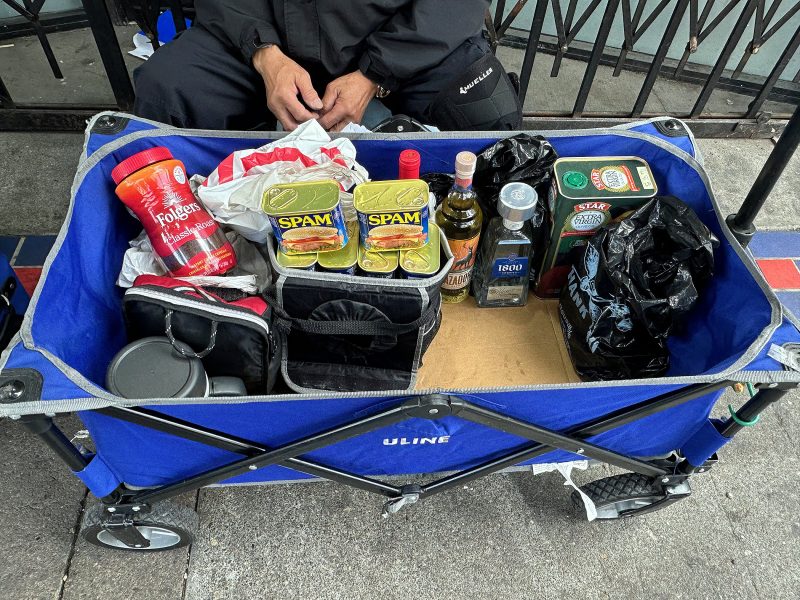 A blue utility cart holds cans of Spam, a jar of Folgers coffee, a bottle of olive oil, alcohol bottles, a thermos, and bags, with a person seated behind it.