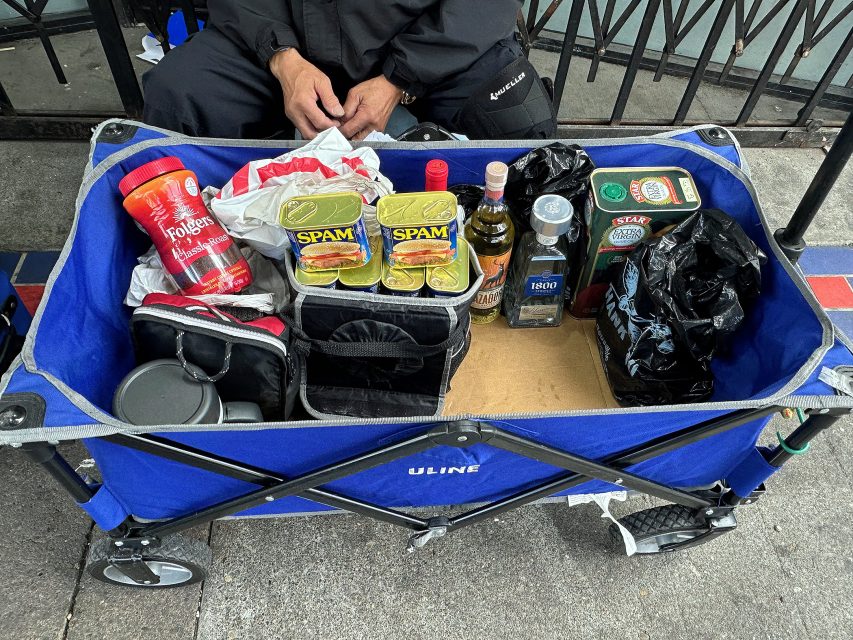 A blue utility cart holds cans of Spam, a jar of Folgers coffee, a bottle of olive oil, alcohol bottles, a thermos, and bags, with a person seated behind it.