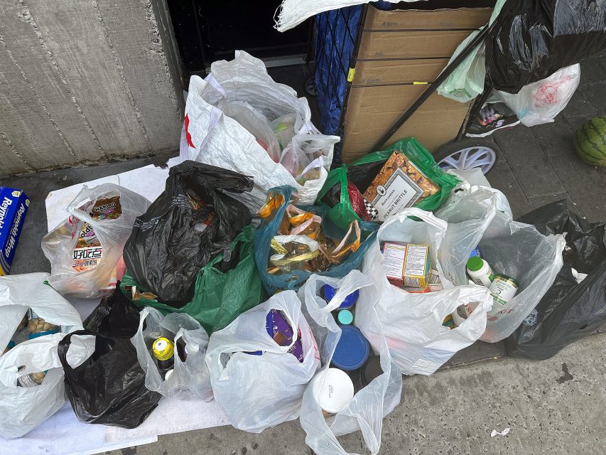 Several plastic bags filled with assorted groceries, including bottles, boxes, and packaged foods, are laid out on a sidewalk next to a wall and a stack of cardboard.