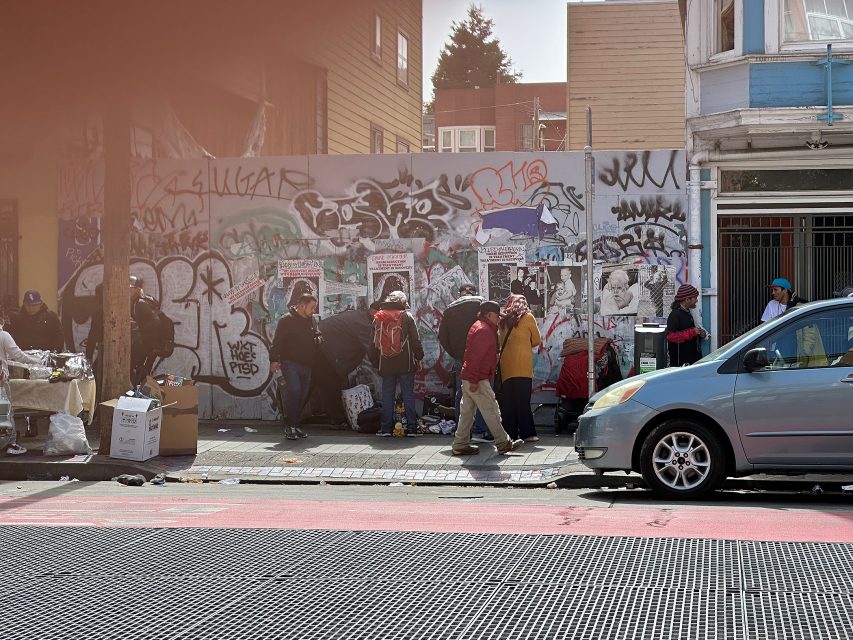 A group of people stand and sit along a graffiti-covered wall on a city sidewalk, with various belongings and tents. A car is parked on the street in front.